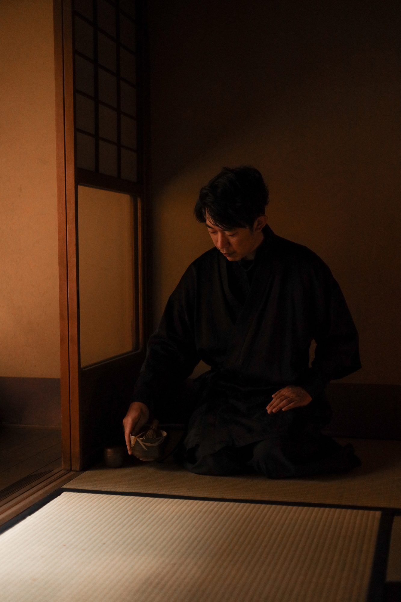 A host kneeling in silence in a dim tea room